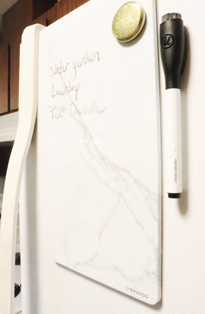 A white magnetic whiteboard with a gold magnet and black dry erase pen attached to the front of a white refrigerator