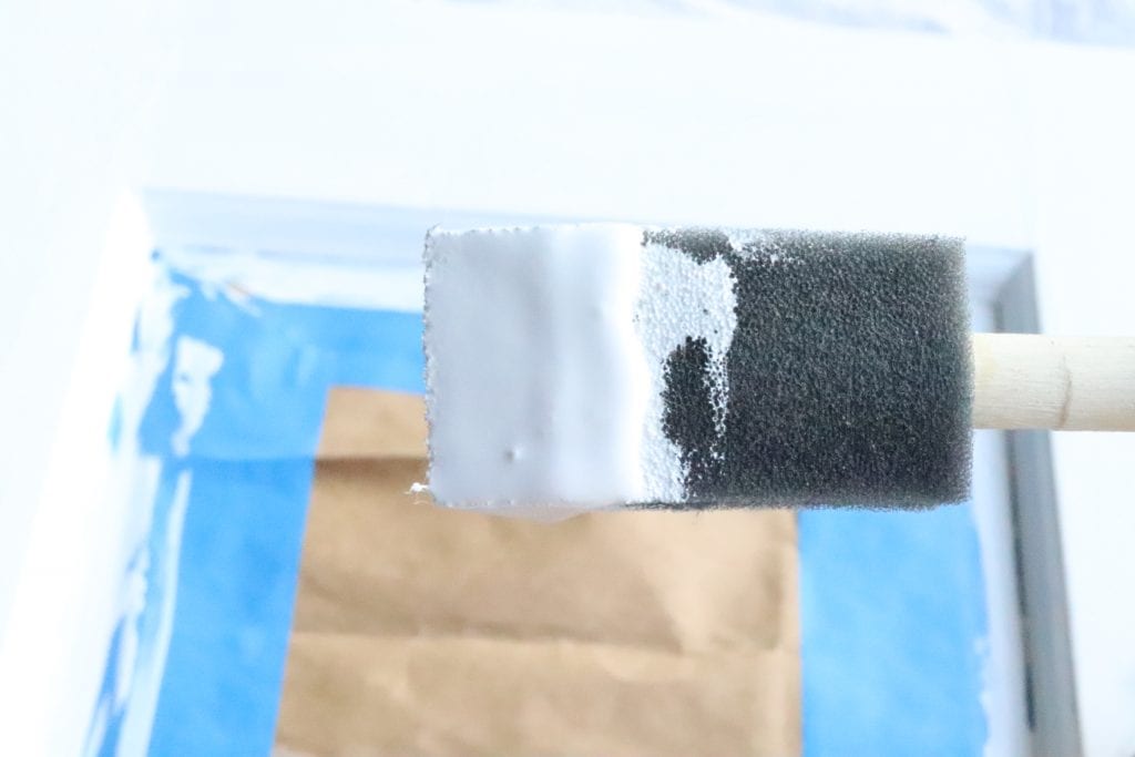 Close-up of a foam brush with white chalk paint on it. Painters tape and brown masking paper in the background.