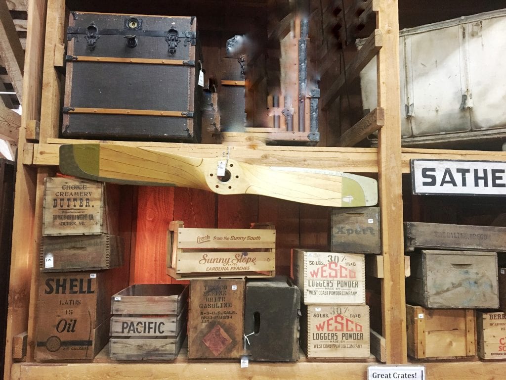 Open shelves of antiques filled with crates and a wooden airplane propellor hanging above one shelf