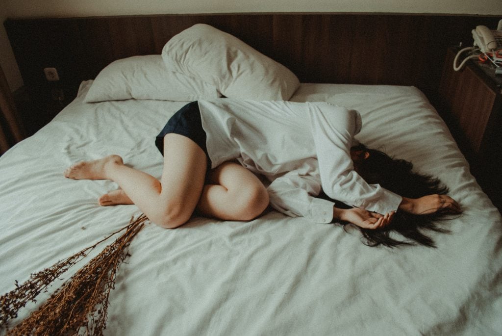 brunette woman in a white blouse and navy shorts lying on a bed with white sheets on it