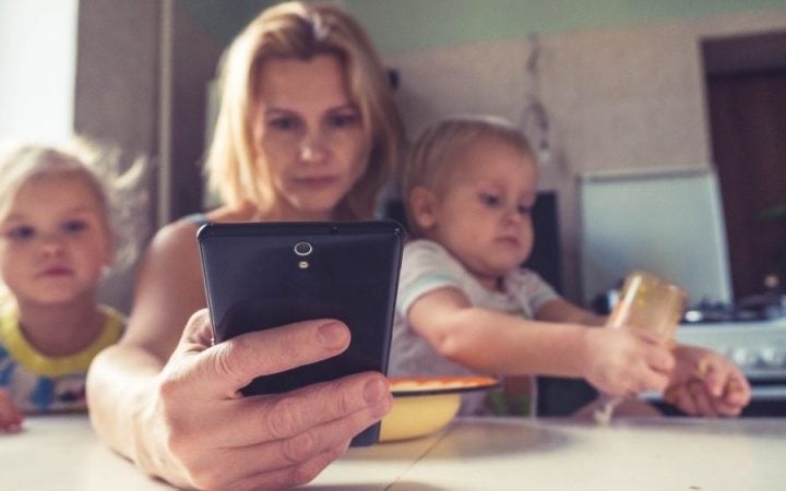 Blonde woman looking at a cell phone while holding a baby and a blonde child at a kitchen table